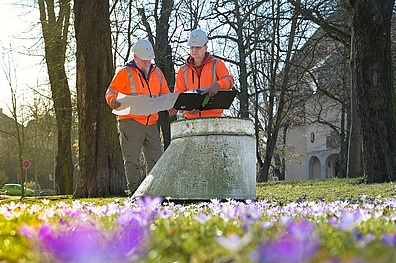 Zwei Männer in orangefarbenen Warnwesten und weißen Schutzhelmen stehen in einem Park und begutachten gemeinsam Pläne bzw. Unterlagen.