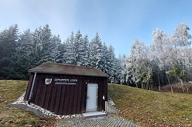 Kleines braunes Holzgebäude mit Schild der Stadtwerke Rehau Hochbehälter Schönlind auf einer gewölbten Wiese, im Hintergrund frost- und schneebedeckte Nadelbäume und Laubbäume bei klarem Winterwetter