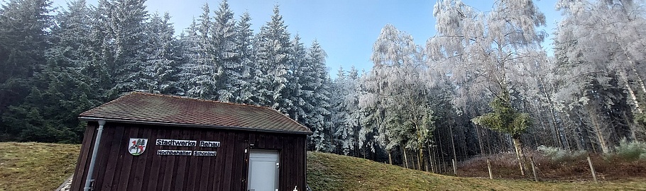 Kleines braunes Holzgebäude mit Schild der Stadtwerke Rehau Hochbehälter Schönlind auf einer gewölbten Wiese, im Hintergrund frost- und schneebedeckte Nadelbäume und Laubbäume bei klarem Winterwetter