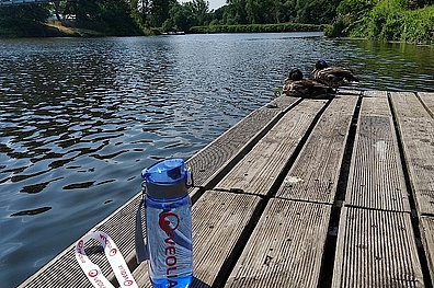Nahaufnahme eines Stegs am Ufer eines Flusses. Auf dem Steg sind zwei Enten zu sehen, eine Veolia Wassertrinkflasche und einer Medaille. Der Fluss im Hintergrund und die natürliche Umgebung vermitteln eine ruhige, idyllische Atmosphäre.