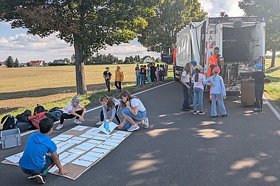 Draußen auf einer Straße neben einem Feld steht im Hintergrund rechts ein Veolia-Müllfahrzeug, daneben eine Gruppe von Grundschulkindern. Ein Mitarbeiter erklärt den Kindern das Fahrzeug. Im Vordergrund sitzen vier Kinder auf dem Boden und spielen ein Memory-Spiel.