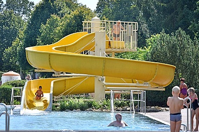Die große gelbe Wasserrutsche vom Muldentalbad Kleinbothen. Die Rutsche hat zwei Ebenen mit geschwungenen Bahnen. Am unteren Ende mündet sie in ein Wasserbecken. Im Hintergrund sind Bäume und ein Gebäude zu sehen.
