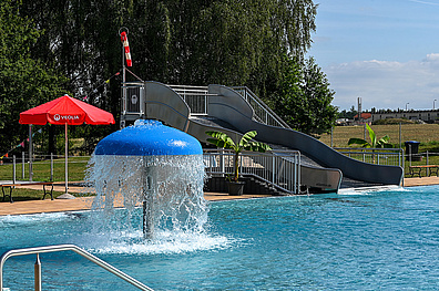 Ein Schwimmbecken mit einer grauen Wasserrutsche und einem blauen Wasserpilz, der Wasser sprüht. Am Beckenrand steht ein roter Sonnenschirm und einige Palmen in Kübeln sorgen für tropisches Flair.