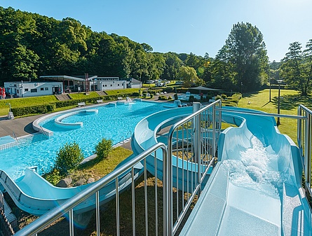 Das Bild wurde auf der Wasserrutsche aufgenommen: Im Zentrum des Bildes ist das große, blaue Schwimmbecken zu sehen, das von weitläufigen grünen Liegewiesen umgeben ist.