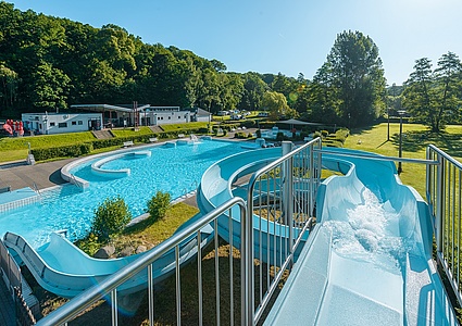 Das Bild wurde auf der Wasserrutsche aufgenommen: Im Zentrum des Bildes ist das große, blaue Schwimmbecken zu sehen, das von weitläufigen grünen Liegewiesen umgeben ist.