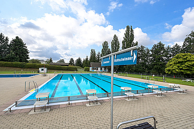 Freibad mit mehreren Schwimmbahnen, Startblöcken und Sprungbrettern. Beschilderung für Schwimmer- und Nichtschwimmerbereiche, umgeben von Bäumen.