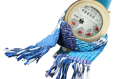 Ein runder, metallischer Wasserzähler mit mehreren Zifferblättern zur Anzeige des Wasserverbrauchs in Kubikmetern. Um den Zähler ist ein blau-weißer Strickschal gewickelt. Dies symbolisiert humorvoll die Gefahr des Einfrierens eines Wasserzählers bei sehr kaltem Wetter.