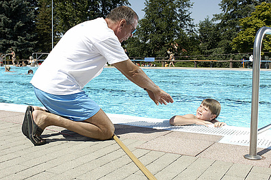 An einem Beckenrand kniet ein Mann in weißem T-Shirt und blauen Shorts. Er beugt sich über den Beckenrand und reicht einem Kind im Wasser die Hand. Das Kind lehnt mit dem rechten Ellenbogen auf dem Beckenrand.