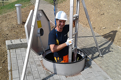 Ein Mitarbeiter in Schutzkleidung und einem Schutzhelm, steht in einem Schacht. Er steht auf einr Leiter die von einem Dreibein-Gestell gehalten wird. Im Hintergrund ist eine sandige Böschung zu sehen.