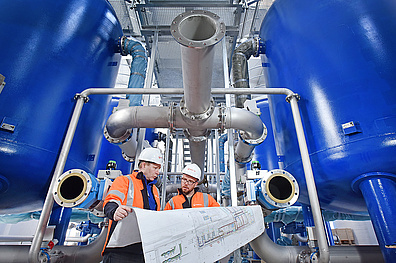 Innenansicht eines Wasserwerks, große blaue Behälter mit vielen Rohren. Zwei Mitarbeiter von Veolia stehen davor und haben einen großen Bauplan in der Hand.