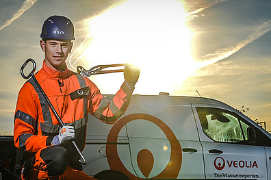 Ein junger Mitarbeiter in auffälliger Arbeitskleidung steht im Vordergrund. In seiner einen Hand hält er einen Kanaldeckelheber, in der anderen eine Rohrzange. Hinter ihm ist ein Fahrzeug von Veolia zu sehen. Im Hintergrund erstrahlt ein greller Sonnenaufgang und taucht die Szene in warmes Licht.