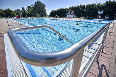 Fotoaufnahme vom Edelstahl-Handlauf am Rand des Schwimmbeckens. Im Hintergrund erstreckt sich das hellblaue Wasser des Schwimmbeckens, in dem einige Menschen schwimmen.