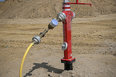 Baustelle mit rotem Hydranten im Vordergrund und gelbem Volvo-Bagger auf einem Sandhaufen im Hintergrund. Ein gelber Schlauch ist am Hydranten angeschlossen.