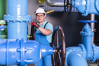 In einem Wasserwerk steht ein Mitarbeiter in Arbeitskleidung zwischen mehreren großen, blauen Wasserrohren. Die Rohre sind mit Drehkreuzen ausgestattet, die zur Regulierung des Wasserflusses dienen.