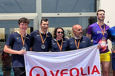 Drei Medaillengewinner stehen auf einem Siegerpodest bei einem Schwimmwettbewerb der Europäischen Betriebssportspiele. Die Athleten in Sportbekleidung präsentieren gemeinsam eine weiße Veolia-Fahne vor dem Hintergrund einer Schwimmhalle.