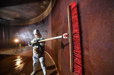 Ein Mitarbeiter in Schutzkleidung bei der Reinigung eines Hochbehälters für Trinkwasser. Er steht in dem großen Behälter und säubert die Innenwände mit einem großen Besen.